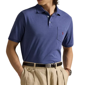 A man wears a blue polo shirt with a pocket featuring a red logo, paired with beige pleated trousers and a black belt.