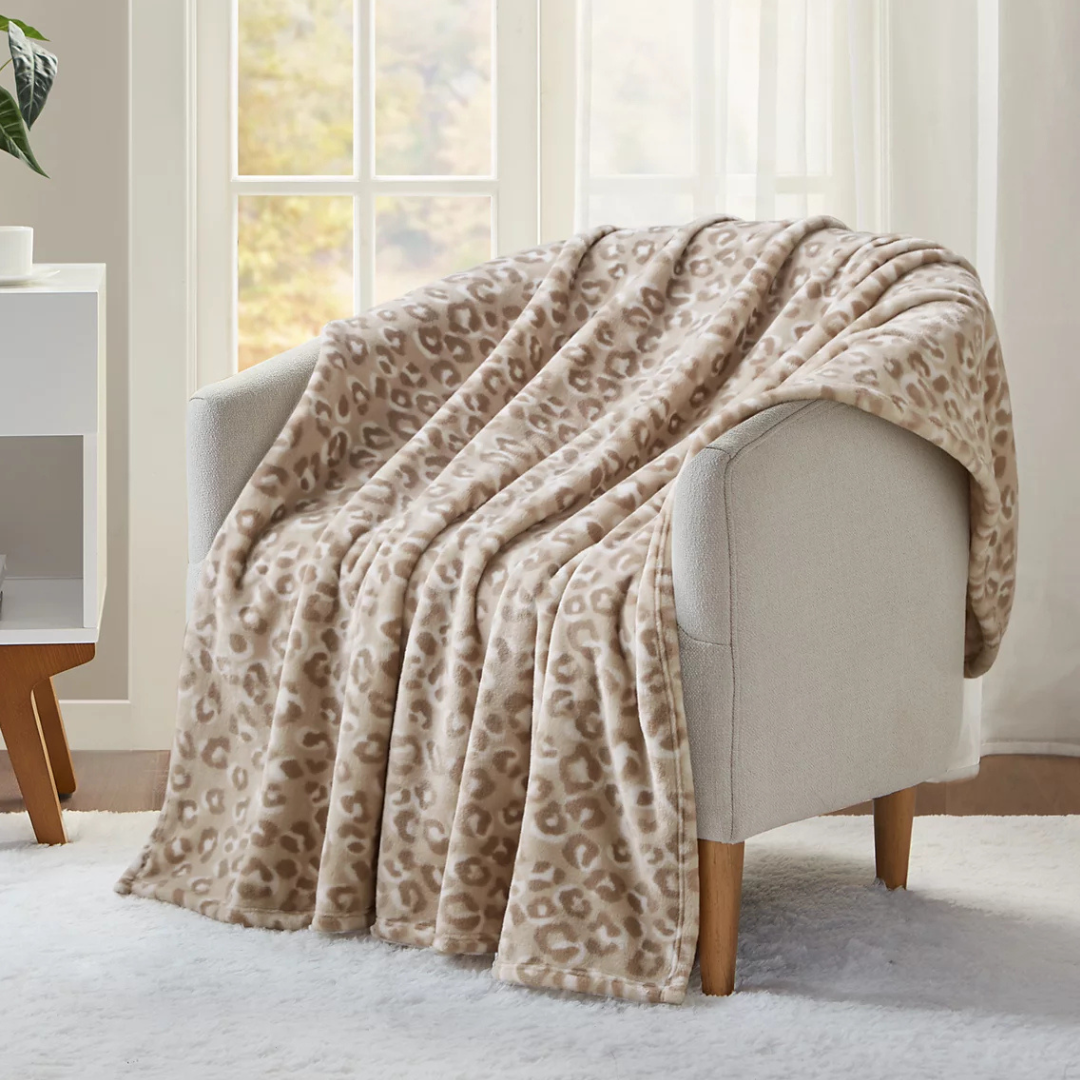 A beige and brown leopard print blanket is draped over a light gray armchair with wooden legs, placed in front of a window.