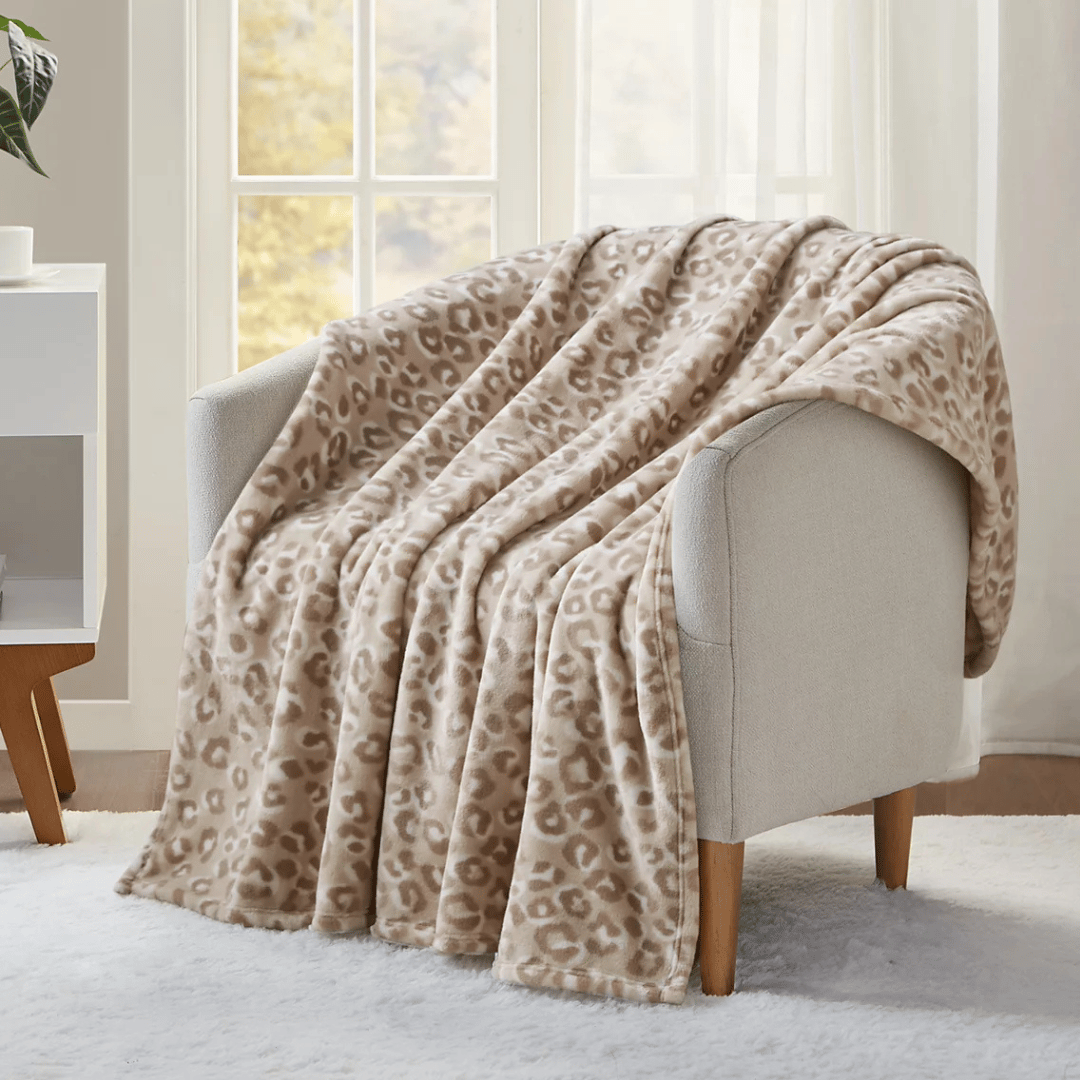 A beige and brown leopard print blanket is draped over a light gray armchair with wooden legs, placed in front of a window.
