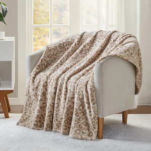 A beige and brown leopard print blanket is draped over a light gray armchair with wooden legs, placed in front of a window.