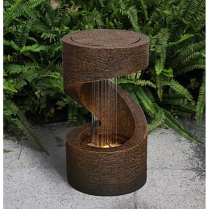 A brown spiral-shaped outdoor water fountain with water cascading down from the top level to a basin below, set against a background of green ferns.