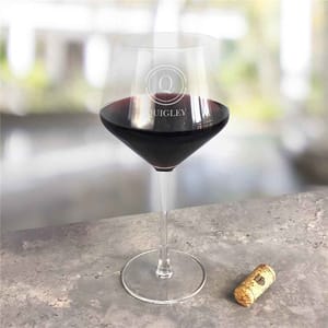 A red wine glass with the name \“Quigley\“ etched on it. The glass is filled with red wine and is placed on a stone surface next to a wine cork.