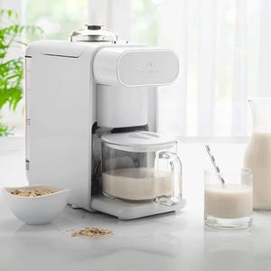 A ChefWave Non-Dairy Milk Maker is present with a sleek white design; it features a glass carafe filled with milk placed beneath the dispenser. Surrounding the appliance are a bowl of oats and a glass of milk, suggesting oat milk preparation.