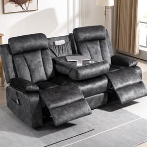 This image features a plush, two-seater reclining sofa in a living room setting. The sofa is upholstered in dark gray fabric and includes a center console with cup holders and a storage compartment, holding a glass and a laptop. Both seats have extendable footrests. The space is styled with a modern lamp, a wooden side table, and a framed artwork on the wall. Neutral curtains allow soft light into the room, enhancing the cozy atmosphere.