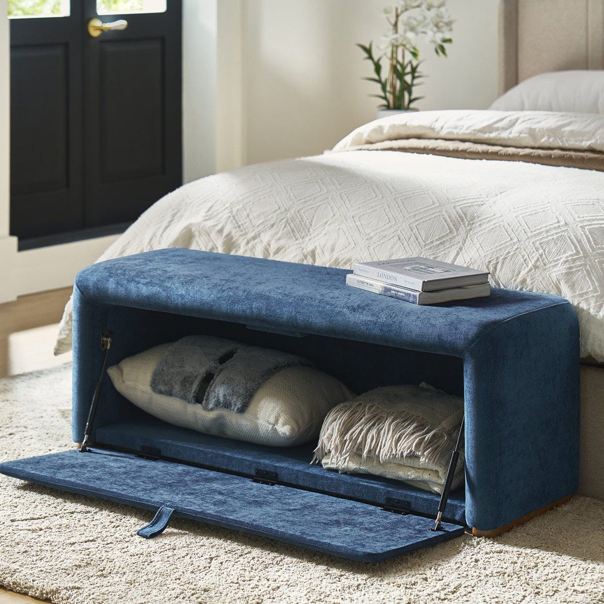 A blue upholstered storage bench with an open compartment containing a pillow and blanket, positioned at the foot of a bed. Books are placed on top of the bench.