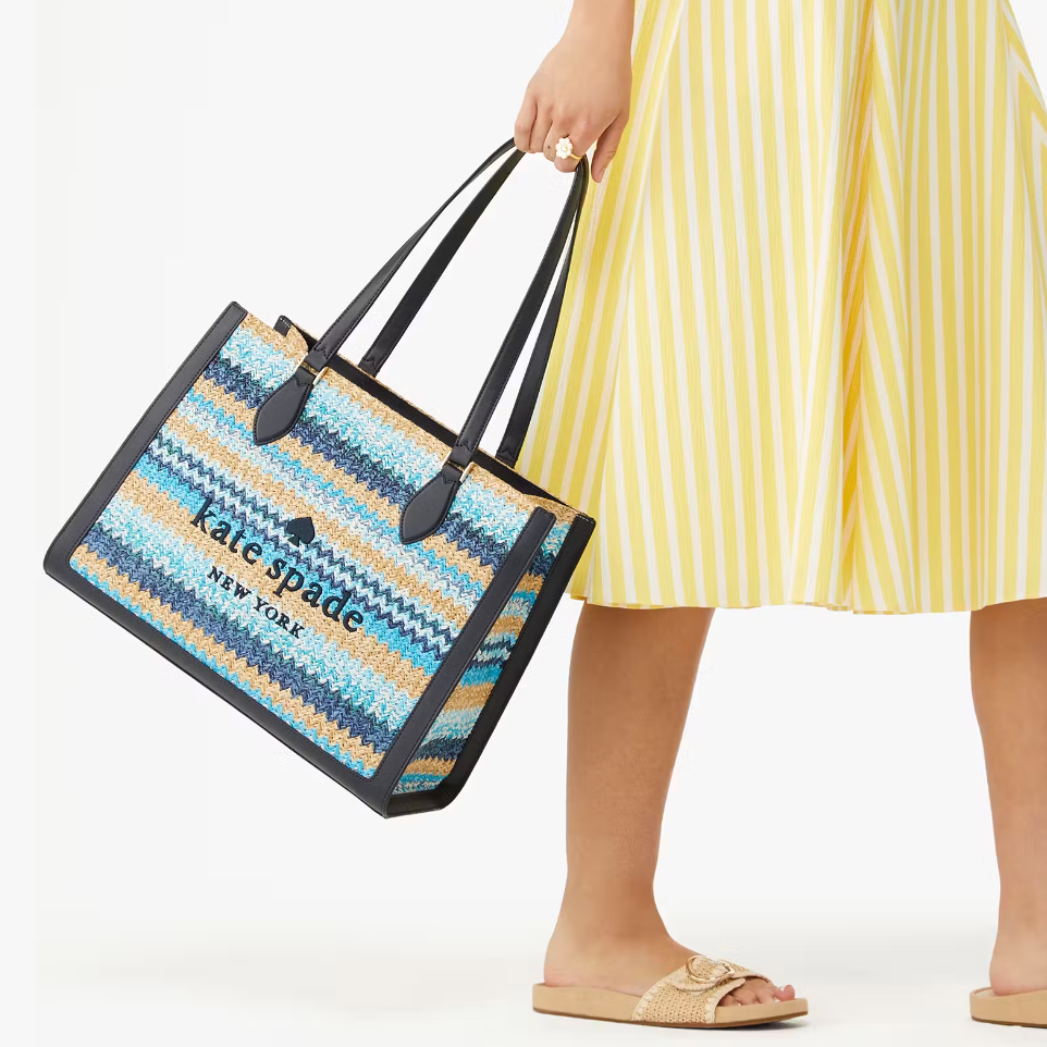 A person is holding a blue and beige striped tote bag with black handles. They're wearing a yellow striped skirt and beige sandals.