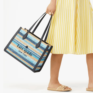 A person is holding a blue and beige striped tote bag with black handles. They're wearing a yellow striped skirt and beige sandals.