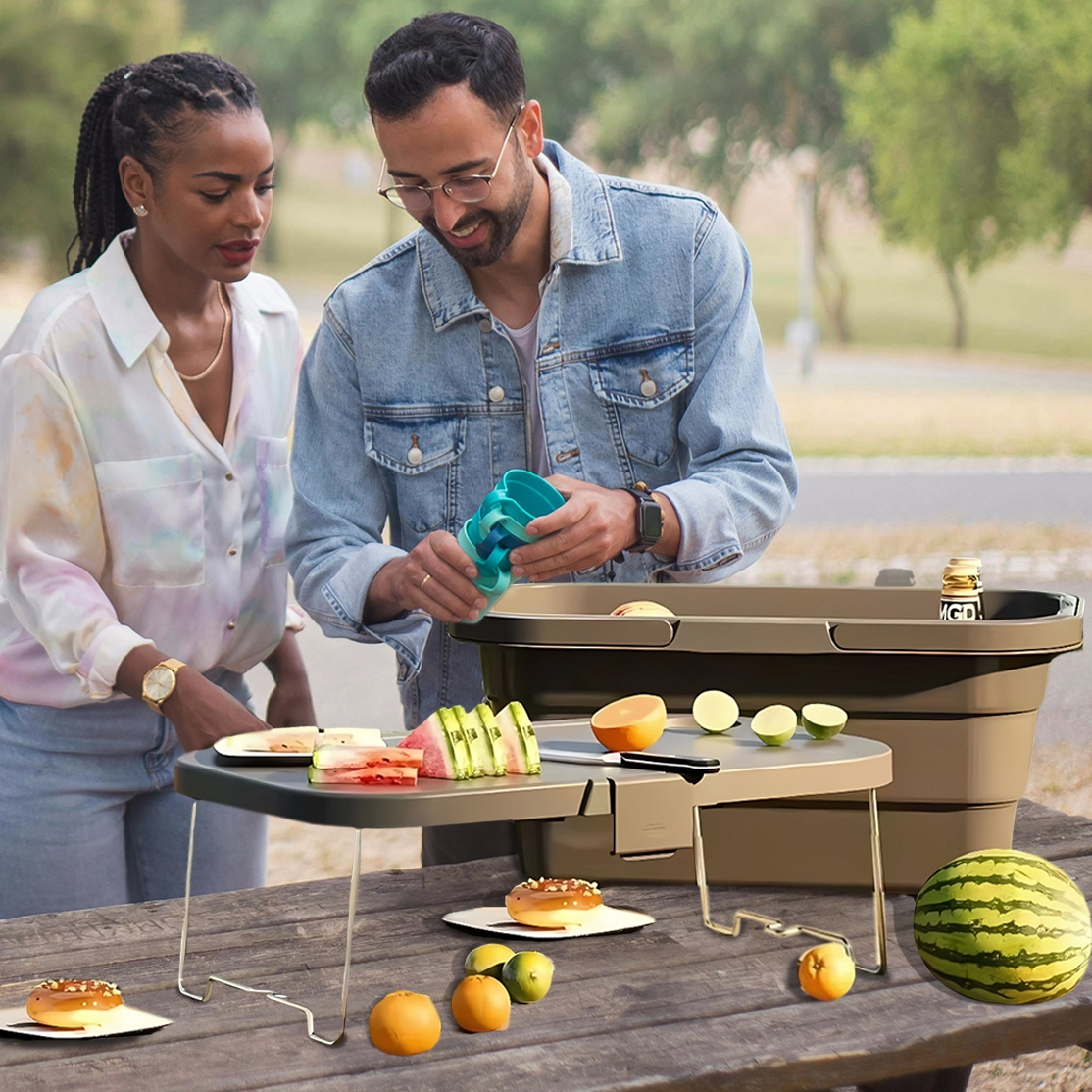 A collapsible picnic basket with a built-in tray featuring foldable legs for stability, ideal for outdoor dining. It includes sections for food and drink storage, with sliced fruits and drinks displayed on and around it.