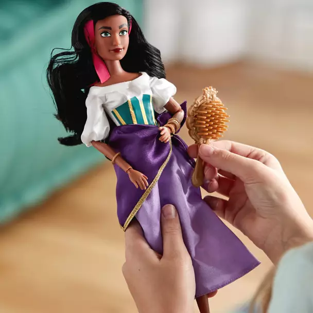 A doll resembling Esmeralda from the \“Hunchback of Notre Dame,\“ with dark hair, a white blouse, and a purple dress, is held by a hand and comes with an accessory resembling a tambourine.