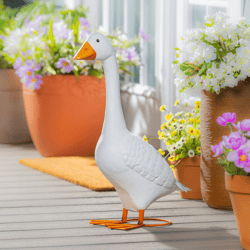 A decorative goose sculpture stands on a porch surrounded by potted flowers with a wooden deck background. The sculpture features a white body and an orange beak and feet.
