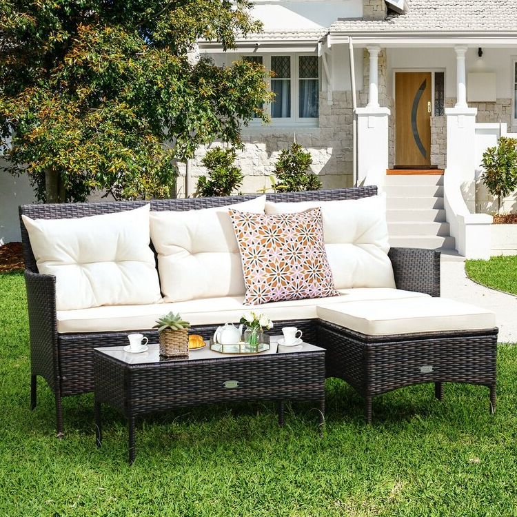 A wicker patio set including a sofa with white cushions, a matching ottoman, and a coffee table, with a decorative pillow and tray with items on the table.