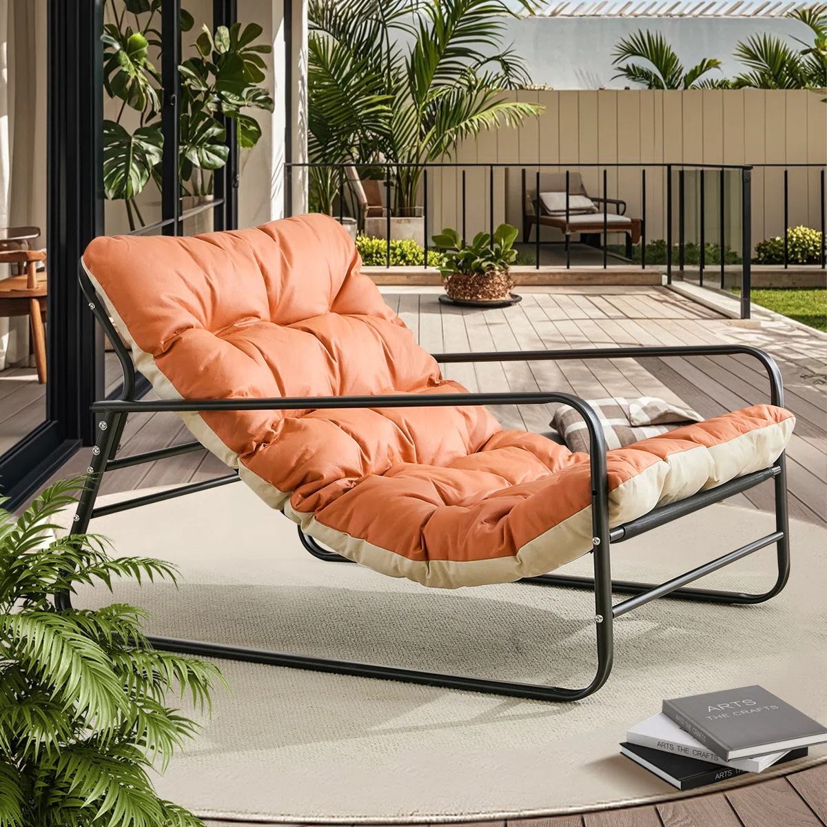 A cushioned lounger with a black metal frame and orange padding is placed on a deck surrounded by greenery, next to a stack of books.