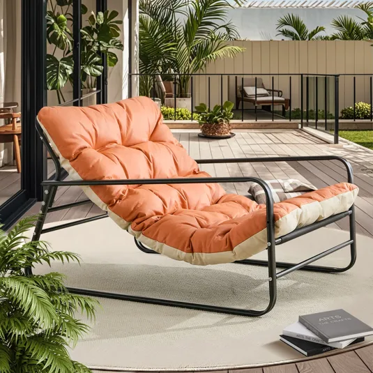 A cushioned lounger with a black metal frame and orange padding is placed on a deck surrounded by greenery, next to a stack of books.