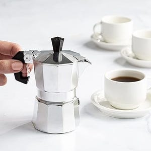 A stovetop espresso maker with a cup of coffee in the background.