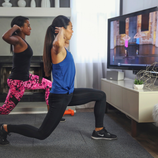 Two people exercise in a living room, following a workout video on a TV. They have dumbbells and are engaged in a lunge position, with one wearing a blue top and the other wearing a black top and pink patterned leggings.