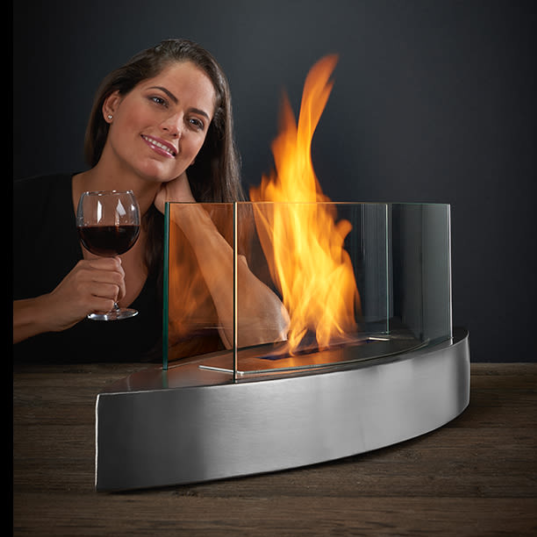 A woman holds a glass of red wine next to a tabletop fireplace with a curved, stainless steel base and glass panels containing the flame.