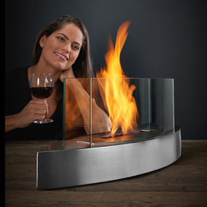 A woman holds a glass of red wine next to a tabletop fireplace with a curved, stainless steel base and glass panels containing the flame.