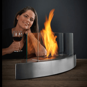 A woman holds a glass of red wine next to a tabletop fireplace with a curved, stainless steel base and glass panels containing the flame.