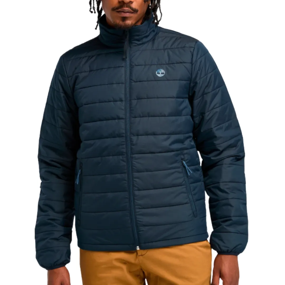 A man is wearing a navy blue, quilted puffer jacket with a high collar and zippered front, featuring a small logo on the chest, paired with mustard-colored pants.