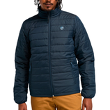 A man is wearing a navy blue, quilted puffer jacket with a high collar and zippered front, featuring a small logo on the chest, paired with mustard-colored pants.