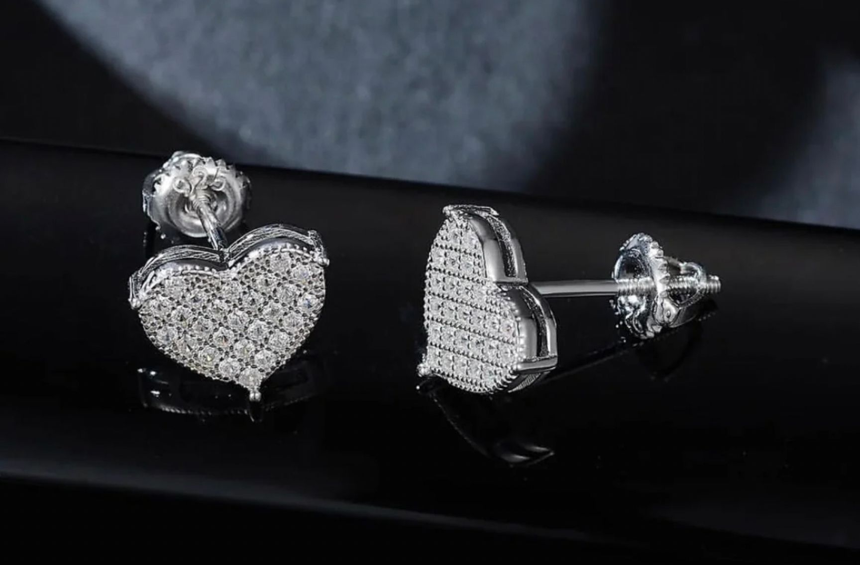 A pair of heart-shaped earrings, each embellished with numerous small moissanite stones set in a reflective silver or white gold metal, showcased against a dark background.
