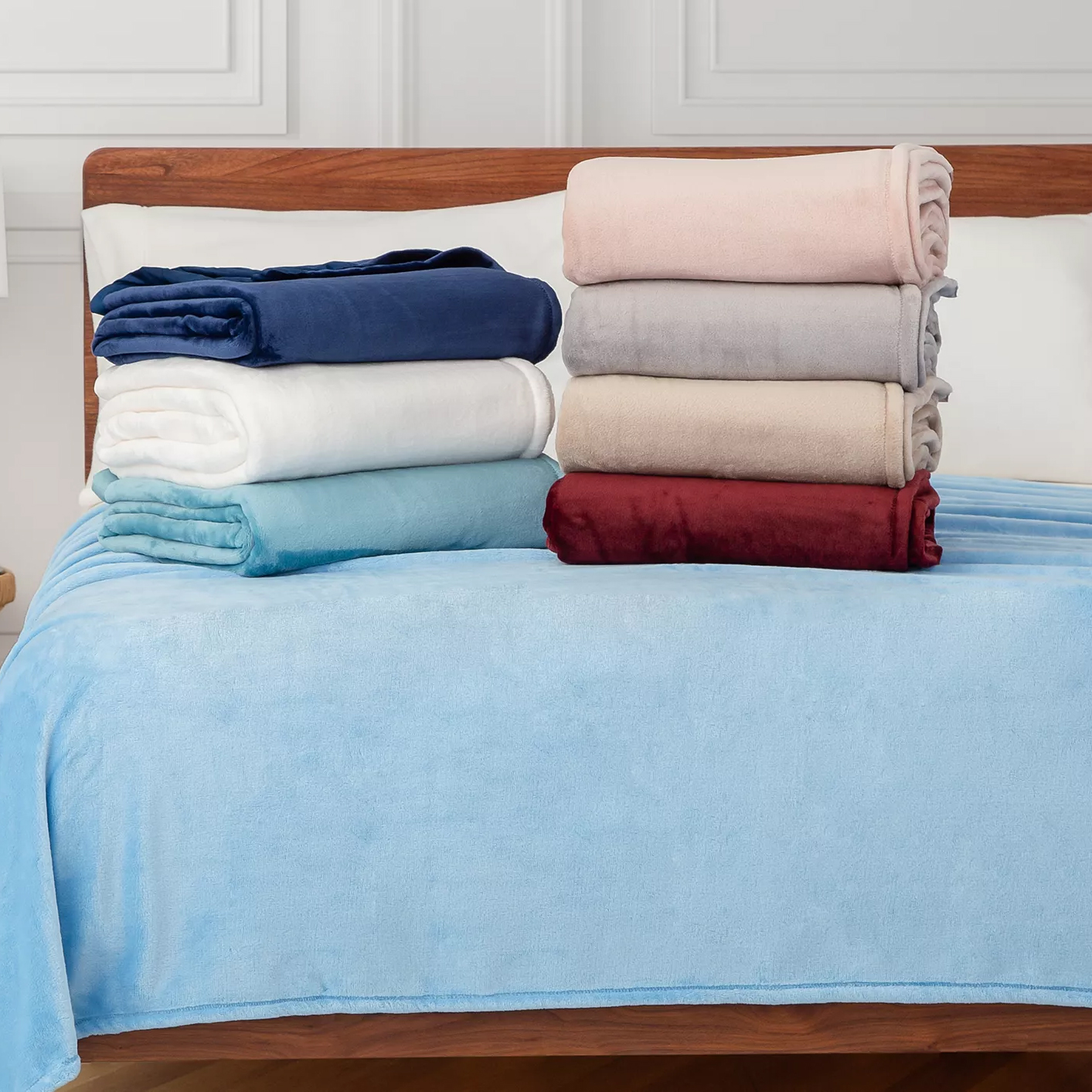 A collection of soft, folded fleece blankets in various colors including navy, white, light blue, pink, gray, beige, and burgundy, displayed on a bed with a light blue blanket spread out.