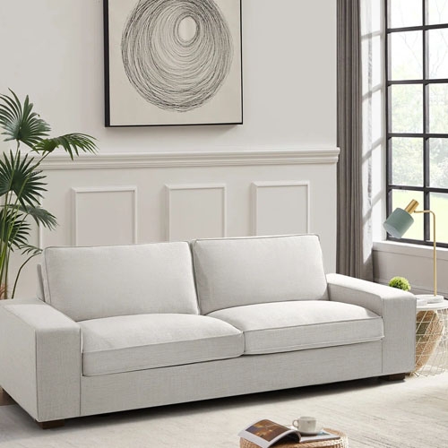 A modern light gray chenille sofa measuring 88.5 inches, featuring clean lines and two large seat cushions. It's set against a white paneled wall with a circular abstract artwork above it. A plant and a window with gray curtains complete the setting.