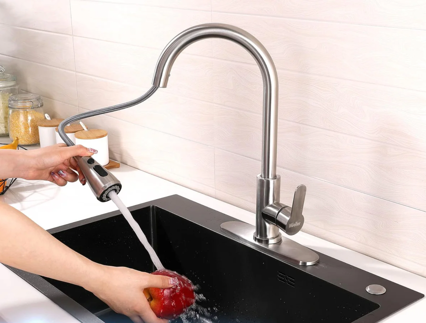 The Mueller Pull-Down Kitchen Faucet features a sleek, stainless steel design with a flexible, retractable hose for easy maneuverability. It includes a single handle for temperature control and is shown being used to rinse an apple in a black sink.