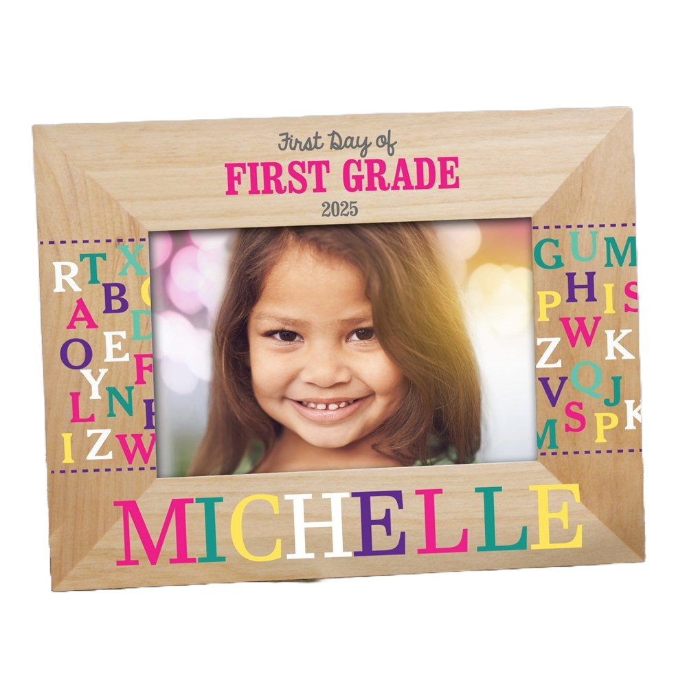 A wooden photo frame with colorful, scattered alphabet letters surrounds a child's photo. \“First Day of First Grade 2025\“ is written at the top, and \“Michelle\“ is in multicolored text at the bottom.