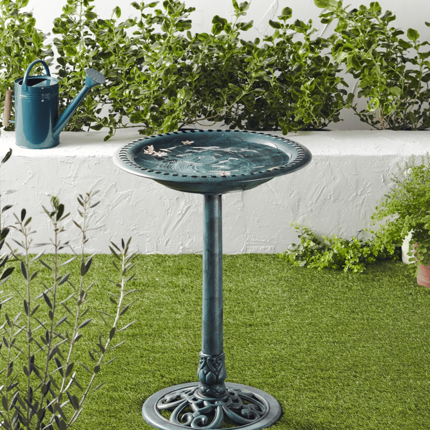 A green birdbath on a pedestal is surrounded by plants and a watering can, promoting a garden's ecological benefits like pest control and soil fertilization.