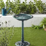 A green birdbath on a pedestal is surrounded by plants and a watering can, promoting a garden's ecological benefits like pest control and soil fertilization.
