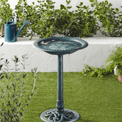 A green birdbath on a pedestal is surrounded by plants and a watering can, promoting a garden's ecological benefits like pest control and soil fertilization.