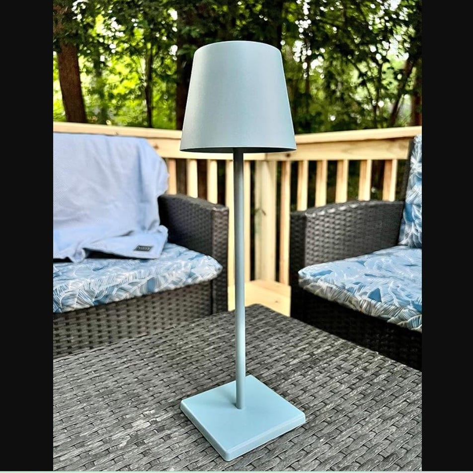 A freestanding lamp with a blueish shade and base, positioned on a wicker outdoor table between two cushioned patio chairs.
