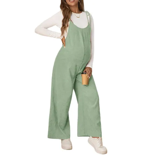 A woman wearing a light green maternity jumpsuit over a white long-sleeve top, holding a takeaway cup and dressed in white sneakers.