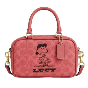 A pink handbag featuring a graphic of a cartoon character named Lucy. It has two handles, a detachable shoulder strap, gold-tone hardware, and a tag.