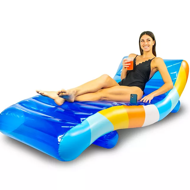 A woman relaxes on a blue and multicolored Chaise Lounge Deluxe Pool Float with built-in cup holder, enjoying a drink in one hand and resting the other on the float.