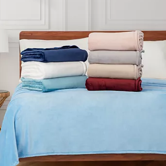 A stack of six folded blankets in various colors is displayed on a bed with a light blue blanket spread across it. The colors include navy blue, white, gray, beige, red, and pink.