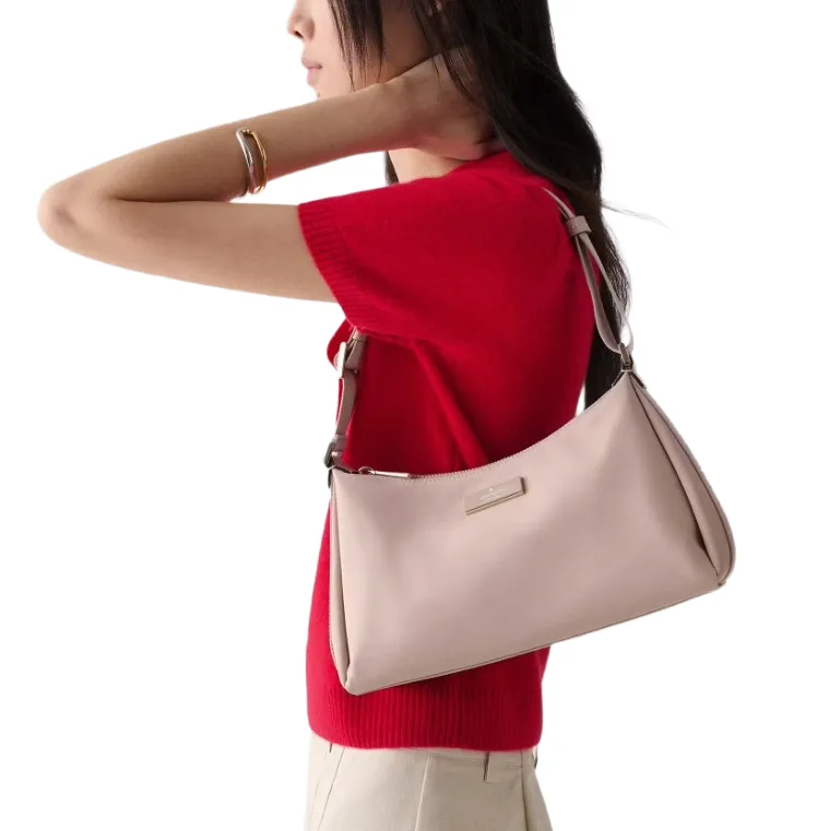 A person wearing a red top and beige pants carries a light pink shoulder bag with a minimalist design and a small logo plaque.