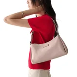 A person wearing a red top and beige pants carries a light pink shoulder bag with a minimalist design and a small logo plaque.