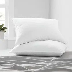 Two white pillows stacked on a neatly made bed with a grey sheet, accompanied by a light-filled room and a plant in the background.