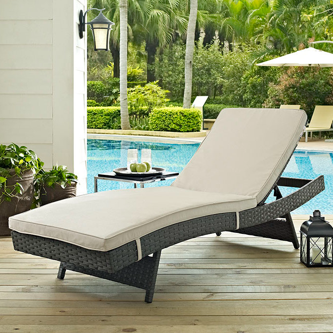 A wicker Sunbrella patio chaise lounger features a beige cushioned seat on a wooden deck next to a pool, surrounded by potted plants, a small table with glasses and apples, and a lantern, with a background of lush green trees and outdoor umbrellas.