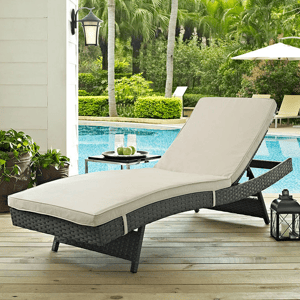A wicker Sunbrella patio chaise lounger features a beige cushioned seat on a wooden deck next to a pool, surrounded by potted plants, a small table with glasses and apples, and a lantern, with a background of lush green trees and outdoor umbrellas.