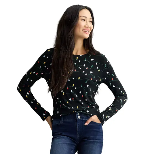 A woman wearing a black long-sleeve shirt with a festive light string pattern and dark blue jeans is standing and smiling.