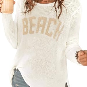 Woman wearing a white knit sweater with \“BEACH\“ text and distressed denim shorts, holding a beverage.