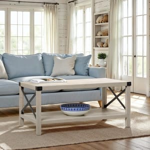 A light wood coffee table with metal accents, featuring a blue and white decorative bowl on the lower shelf. It's placed in a cozy living room with a light blue sofa, soft cushions, and bright natural light from large windows.