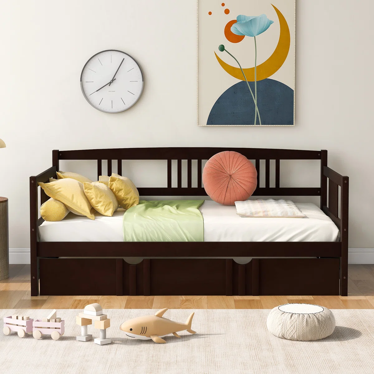 A dark wooden daybed with slatted back and side rails features storage drawers underneath. It is styled with colorful pillows and set against a wall with a clock and abstract art. A rug with wooden toys is placed in front.
