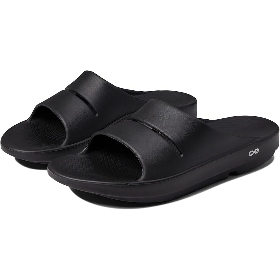 Black slip-on slides with a single wide strap, open toe, and cushioned footbed featuring a slight wedge. The side features a small, circular logo.