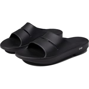 Black slip-on slides with a single wide strap, open toe, and cushioned footbed featuring a slight wedge. The side features a small, circular logo.