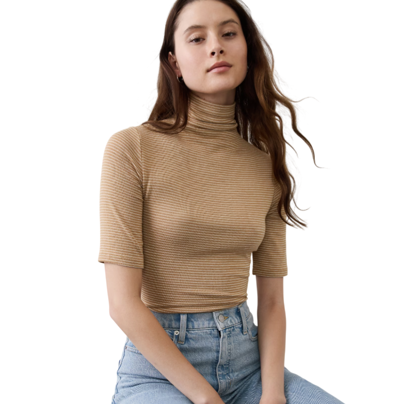 A person wearing a beige striped turtleneck with short sleeves and high-waisted blue jeans seated on a wooden stool against a textured wall.
