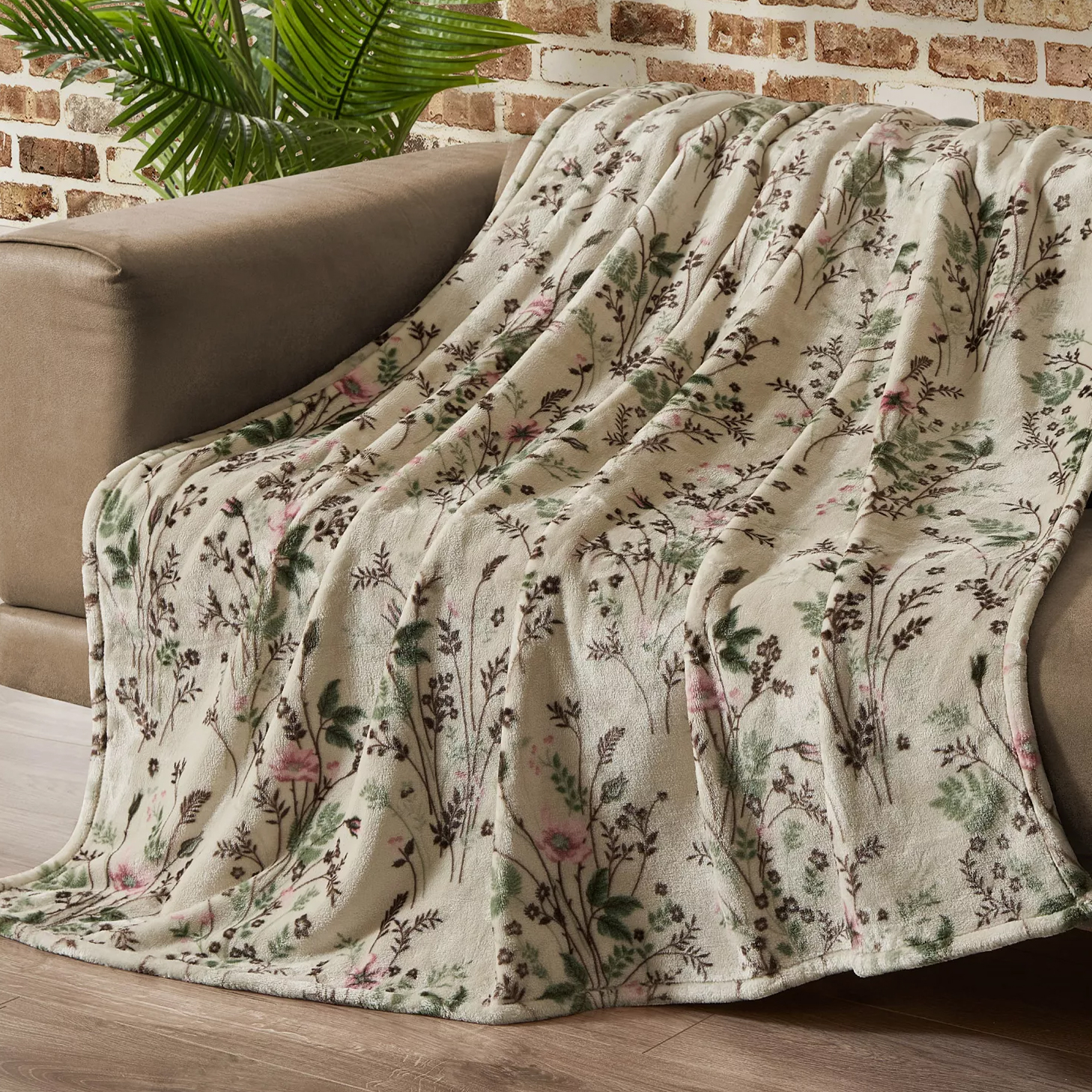 Floral-patterned blanket draped over a brown sofa, set against a brick wall and wooden floor, with a green plant in the background.
